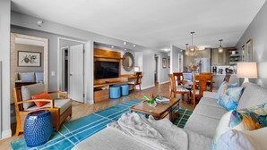 A modern living room with a sofa, coffee table, and TV. There is a dining area with chairs and a table. The kitchen is visible in the background. - Luxury ocean view suite just across street from Waikiki Beach! Free parking! (Honolulu)