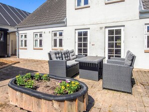Eine Terrasse mit Korbmöbeln, einem Holzpflanzgefäß mit Blumen und einem weißen Gebäude mit Fenstern.