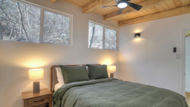 Ein Schlafzimmer mit einem Bett, zwei Nachttischlampen, einem Deckenventilator und großen Fenstern mit Blick auf Bäume.