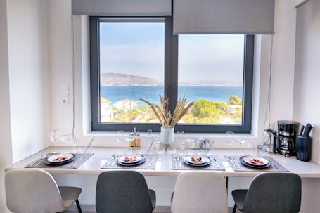 A dining area with a table set for two, a view of the sea, and a window with a view of buildings and hills.