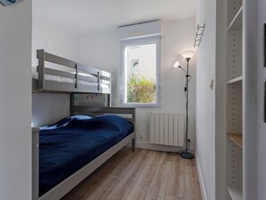 Quarto pequeno e bem iluminado com beliche, janela com vista para vegetação, luminária de chão e radiador.