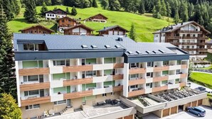 A modern multi-story hotel with balconies, a parking area, and a view of surrounding houses and greenery. - Marmotta by Interhome (Leukerbad)