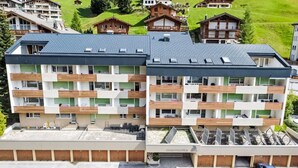 A multi-story hotel with a modern design, featuring balconies, a parking area, and a view of the surrounding green landscape. - Marmotta by Interhome (Leukerbad)
