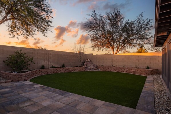 A landscaped backyard with a curved lawn, stone retaining walls, and a variety of trees. - Sleeps 8 | King Suite | Screened Patio + BBQ (San Tan Valley)