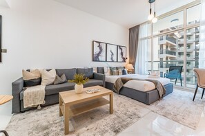 Ein modernes Wohnzimmer mit einem grauen Sofa, einem Holztisch und einem großen Bett mit grauem Kopfteil. An der Wand hängen drei gerahmte Kunstwerke, und ein großes Fenster bietet Blick auf ein Gebäude.