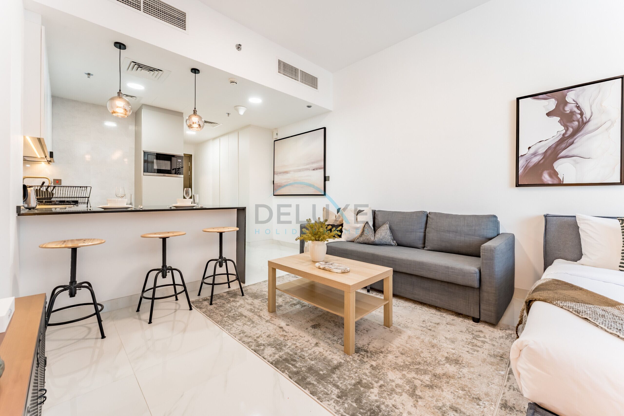 A modern living room with a grey sofa, a wooden coffee table, and three bar stools. There is a kitchen area with a countertop and a sink.