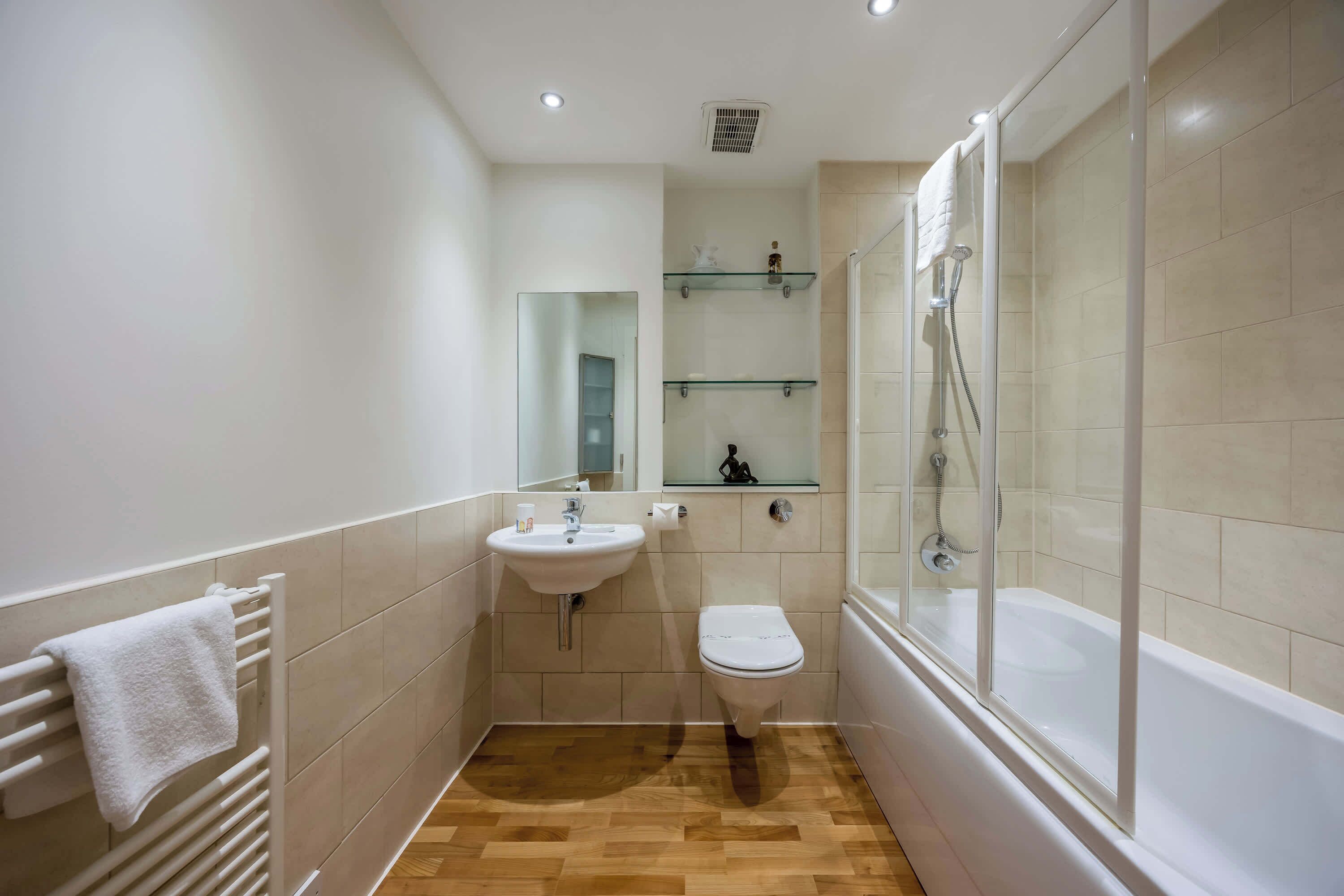 A modern bathroom with a white sink, toilet, and bathtub. There are glass shelves above the sink and a glass shower enclosure.