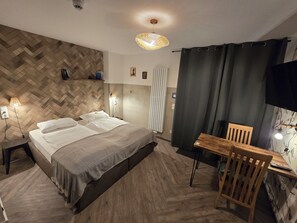 Un dormitorio moderno con un cabecero de diseño geométrico, una cama con ropa blanca, una mesita de noche con lámpara, un escritorio con silla y un televisor montado en la pared.