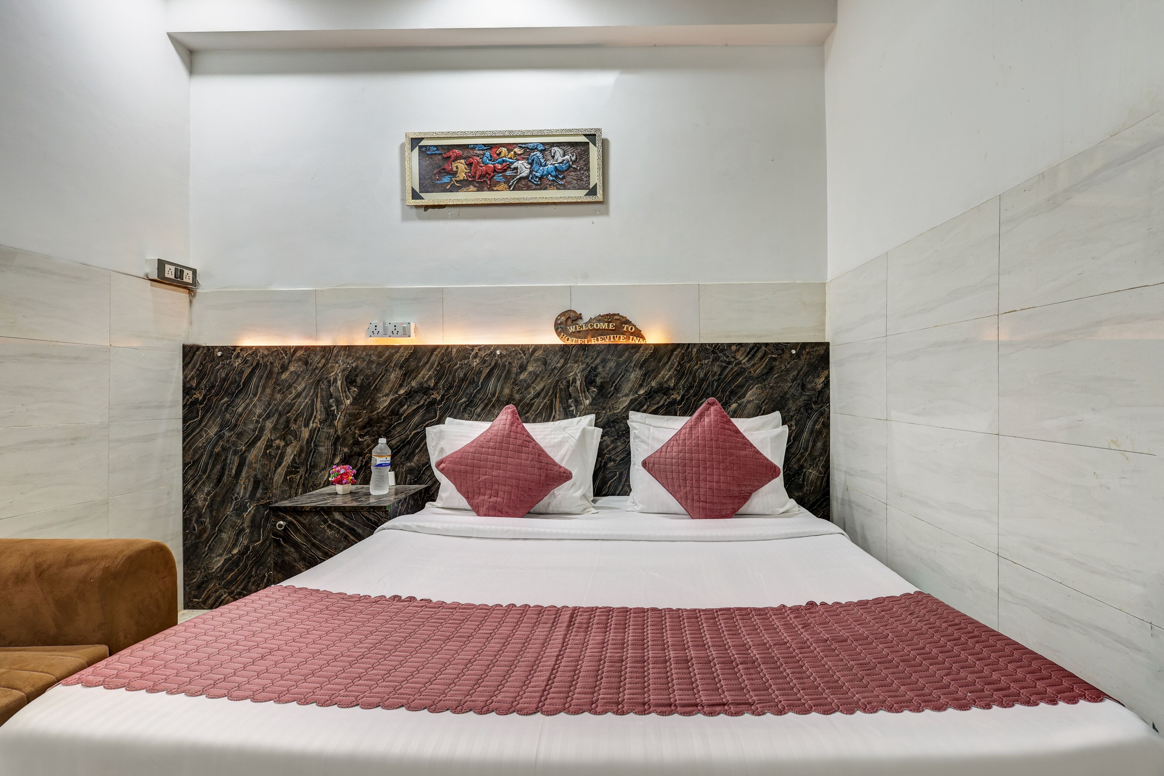 Une chambre d’hôtel avec un lit, une table de chevet, une horloge, un panneau d’accueil et un tableau encadré au mur.
