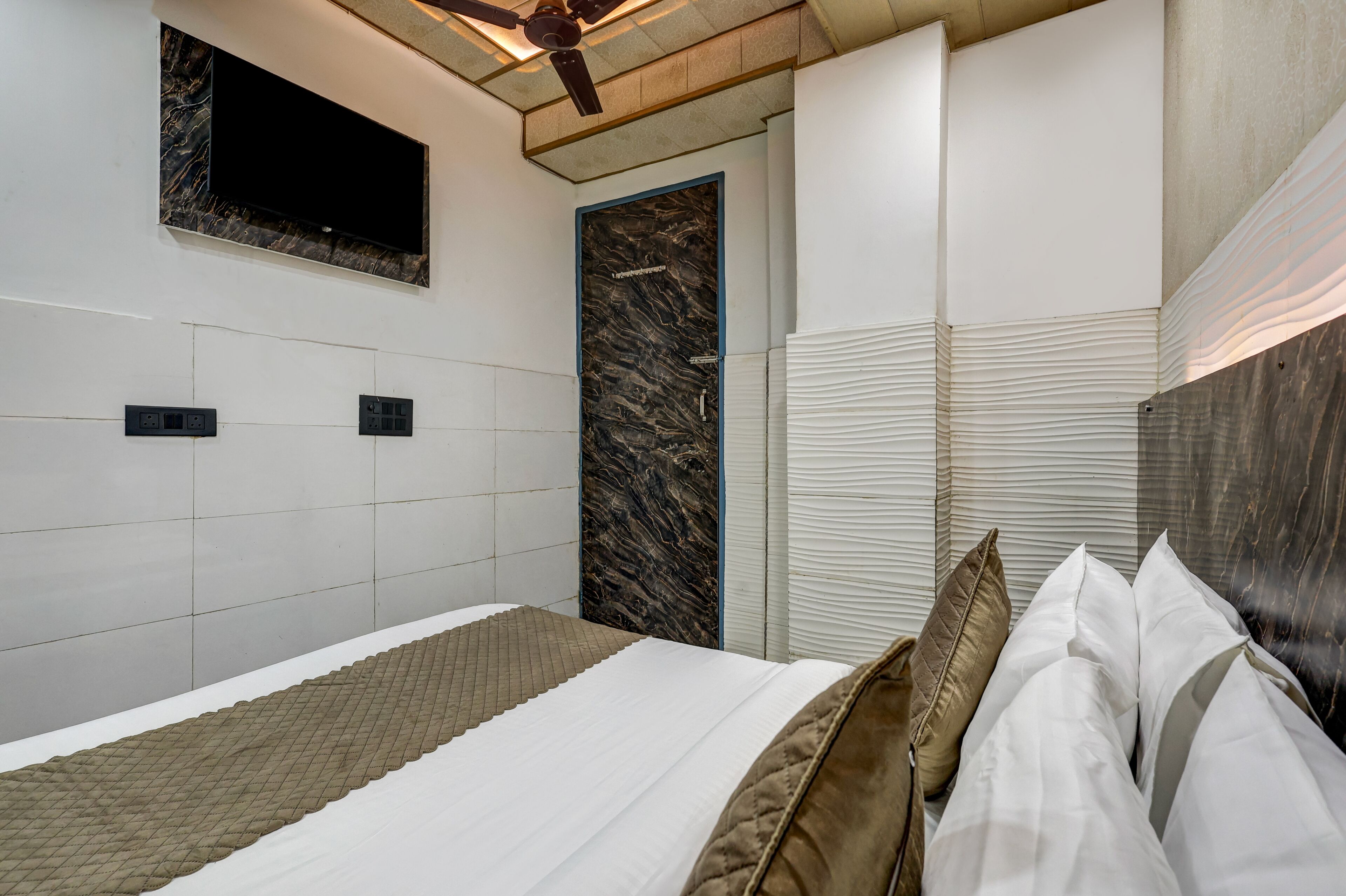 A modern bedroom with a bed, a flat-screen TV mounted on the wall, a ceiling fan, and a door with a marble-like panel.
