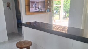 A kitchen with a black countertop, two wooden stools, a TV mounted on the wall, and a view of the outdoors through a window. - Complete House in Pontal do Peró: Comfort and nature next to Búzios (Cabo Frio)