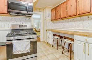 A kitchen with a gas stove, oven, microwave, and wooden cabinets. There are two bar stools and a window with blinds.
