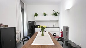 A dining area with a wooden table, chairs, a vase with flowers, a white table runner, a black refrigerator, a sink, and potted plants on the wall. - Guesthouse Delle Navi - Room 3 (Roma)