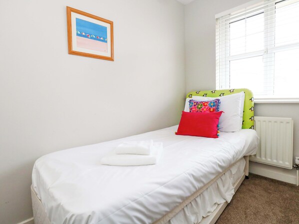 Una cama pequeña y ordenada con ropa de cama blanca y un almohátero rojo, un cuadro en la pared y una ventana con persianas.