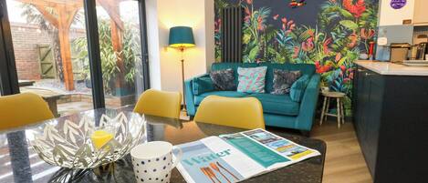 A modern living room with a teal sofa, yellow chairs, a dining table with a glass bowl and muffin, and a kitchen area with a colorful mural.