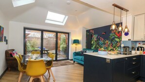 A modern kitchen with a central island, a dining area with yellow chairs, and a view of the garden through a sliding door. - Still Waters (Weymouth)