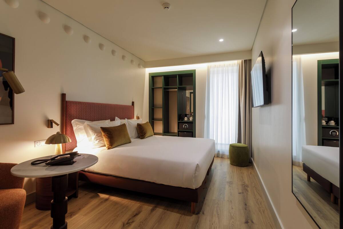A hotel room with a bed, bedside table, lamp, mirror, and a view of the outdoors.