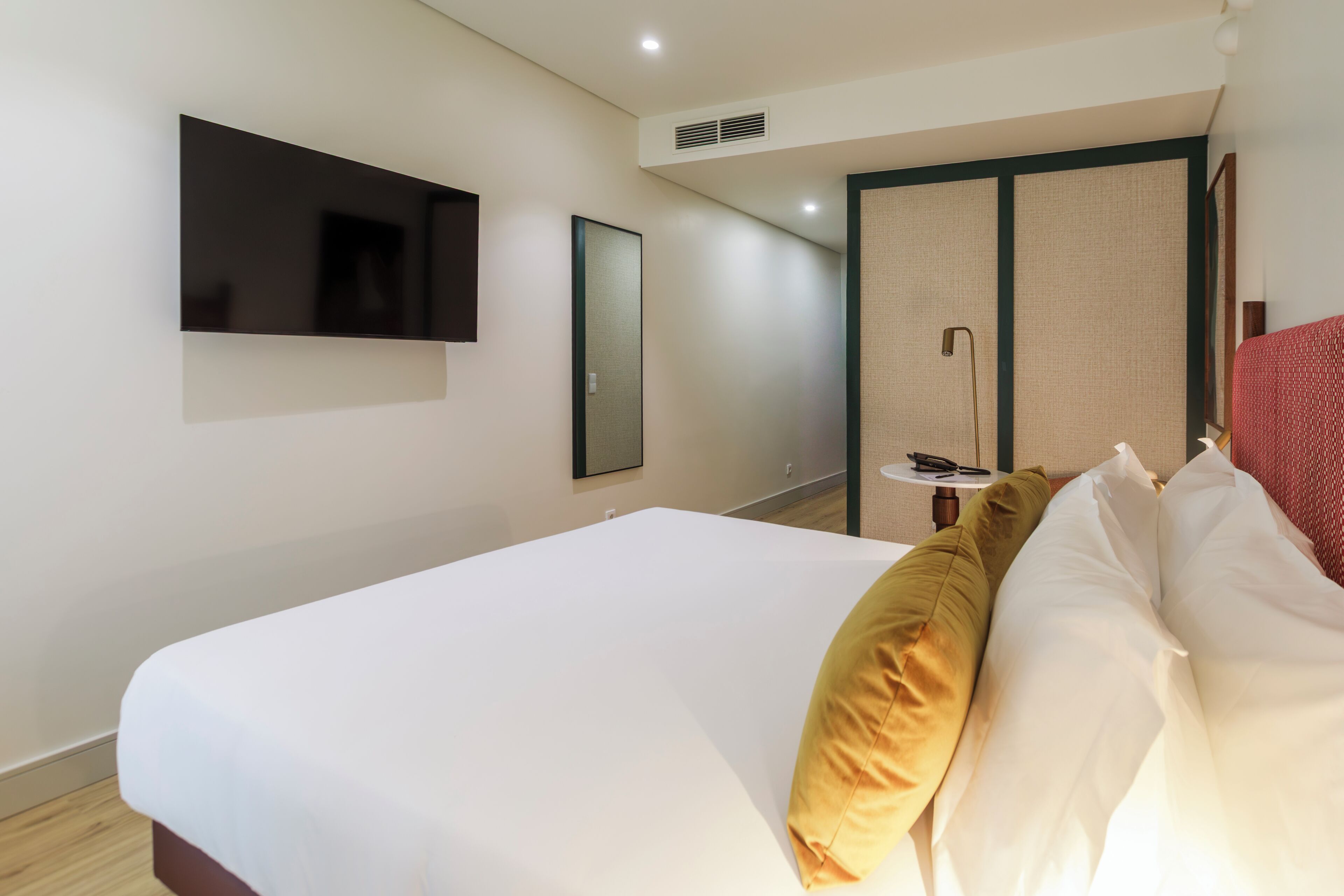 Habitación de hotel con una cama, un televisor en la pared, un espejo y una mesita de noche con una lámpara.