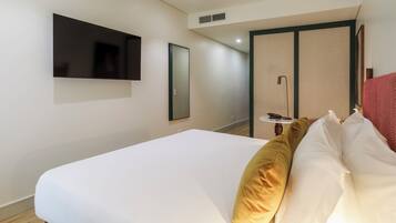 Habitación de hotel con una cama, un televisor en la pared, un espejo y una mesita de noche con una lámpara.