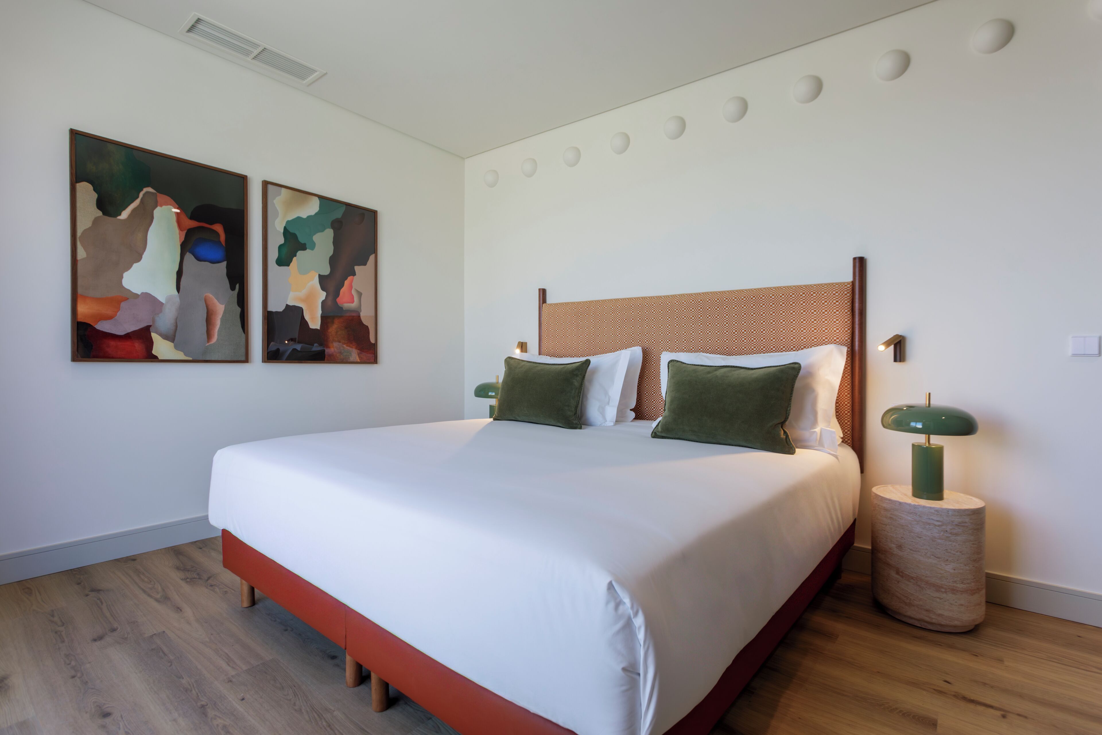 Ein modernes Schlafzimmer mit einem großen Bett, zwei abstrakten Bildern, einem runden Beistelltisch und einer grünen Lampe.