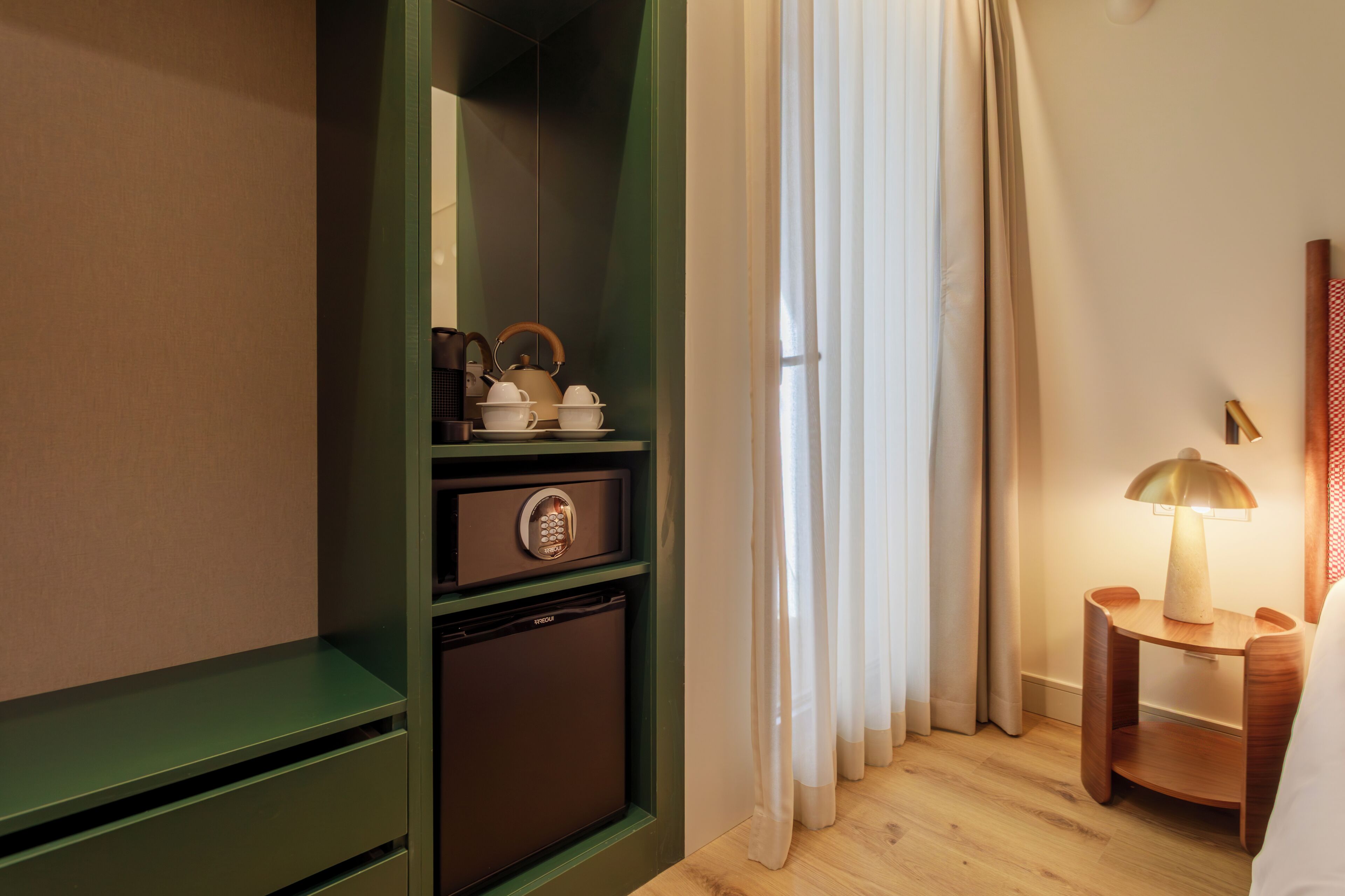 Ein Hotelzimmer mit einem grünen Kleiderschrank, einem kleinen Kühlschrank, einem Nachttisch mit Lampe sowie einem Fenster mit Vorhängen.