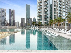 A swimming pool with lounge chairs and palm trees, surrounded by high-rise buildings. - Peninsula 3 - 1320 1BD (Dubai)