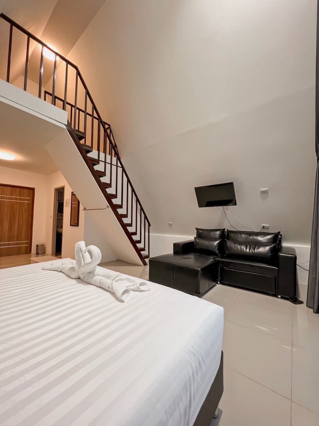 A hotel room with a large bed, a black leather sofa, a flat-screen TV, and a staircase.