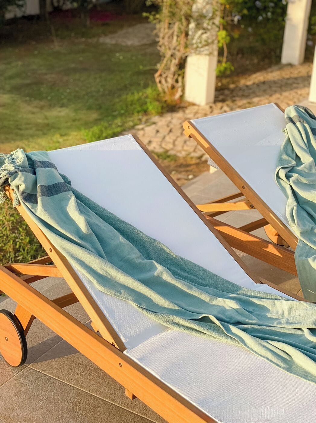 A sun lounger with a light blue towel draped over it, positioned on a patio with a garden in the background.