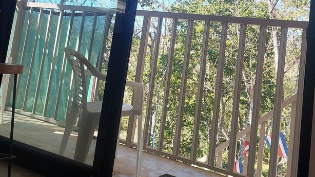 Una stanza con un'ampia porta a vetri scorrevoli che si apre su un balcone esterno con ringhiera bianca e vista su alberi e cielo sereno.