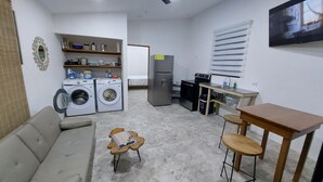 Un ambiente compatto con divano, un piccolo tavolo, lavatrice, frigorifero, forno a microonde e TV a parete.
