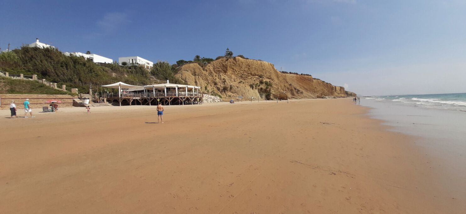 A beach with a white building on a hill, a covered area with tables, and a sandy shore.