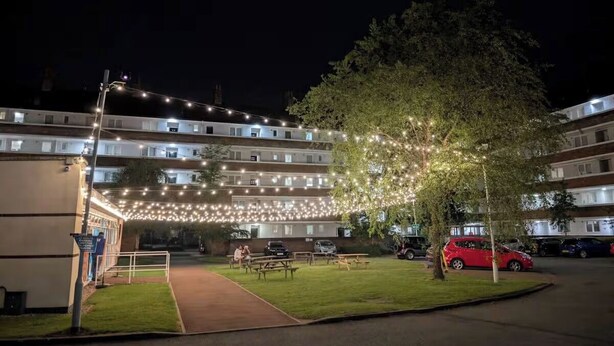 Edificio residencial de varios pisos con un espacio exterior bien iluminado que incluye luces colgantes, mesas de picnic y un árbol.