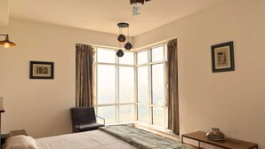 Ein Hotelzimmer mit großem Fenster, einem Bett mit grüner Bettdecke, einem Sessel, einem kleinen Tisch mit Vase und einem Deckenventilator.