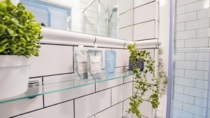 A bathroom with white tiled walls, a glass shelf holding toiletries, and a potted plant. - Charming Suite at the Marina (Howth)