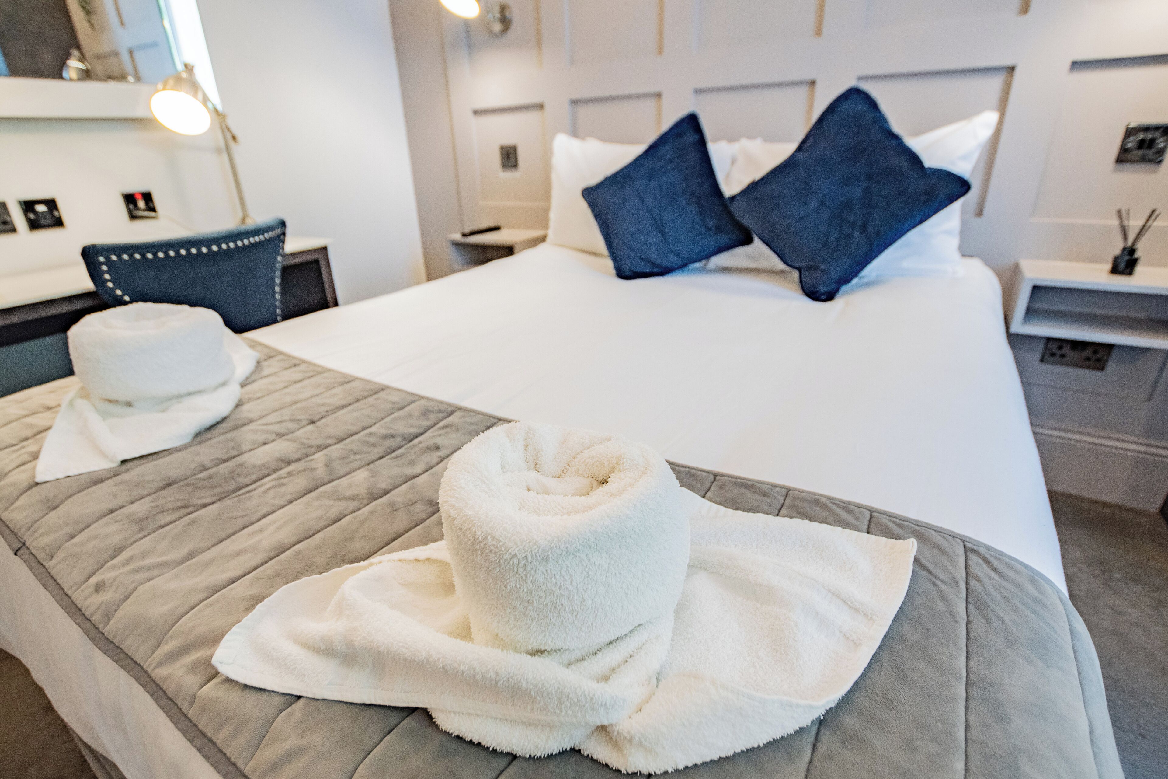A hotel room with a neatly made bed, a chair, a desk, and a nightstand with a lamp.