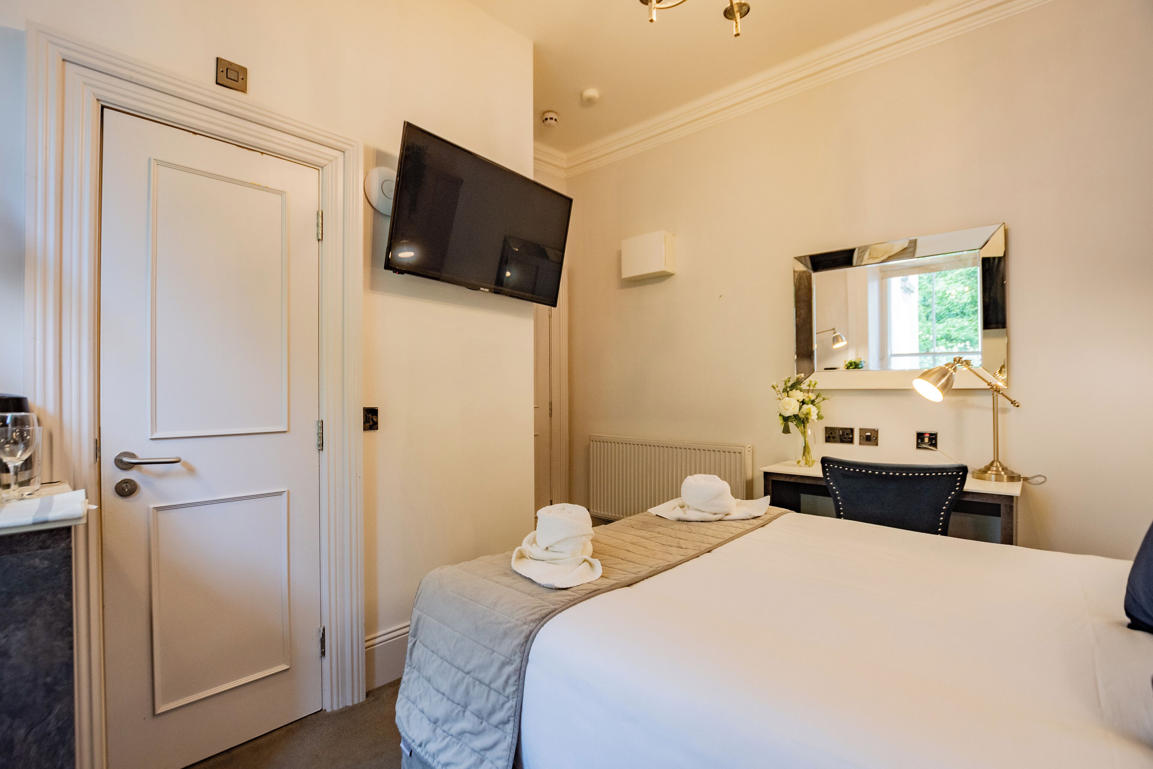 Habitación de hotel con una cama, un escritorio con lámpara, un televisor montado en la pared y un espejo sobre el escritorio.