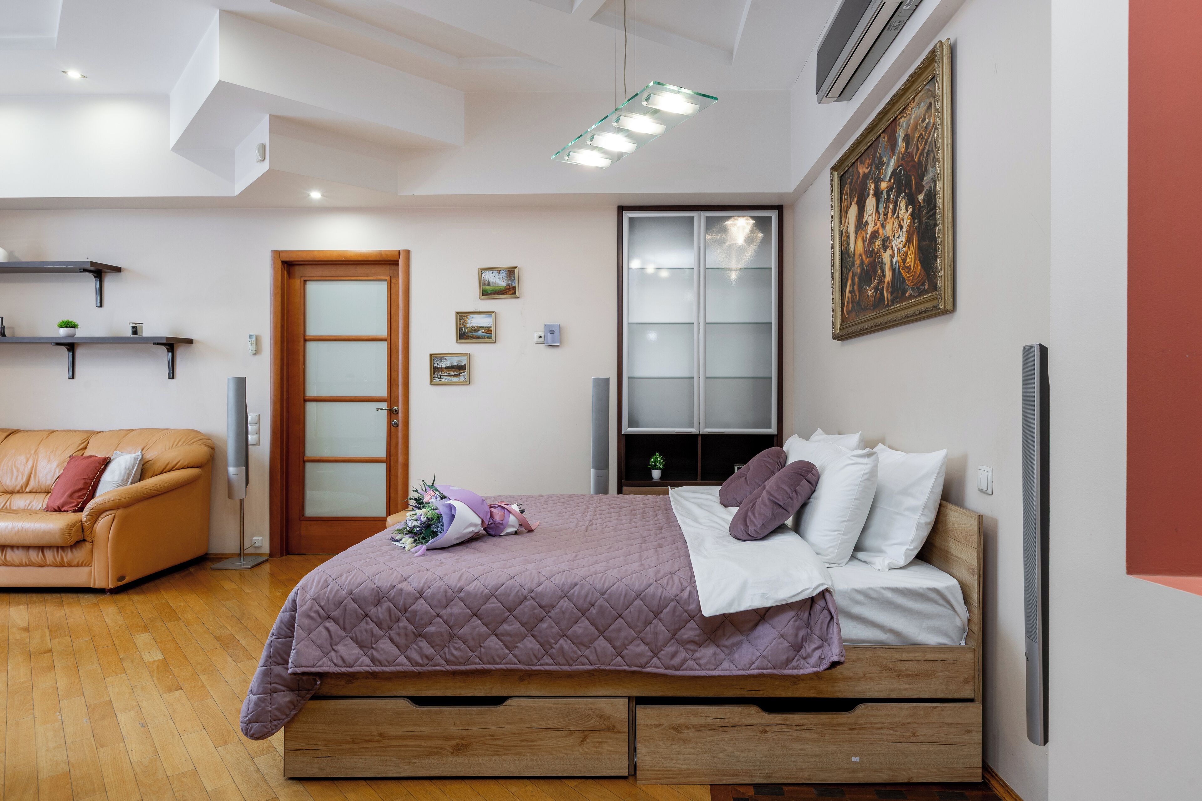 A bedroom with a bed, a wooden floor, a sofa, a door, and a painting on the wall.
