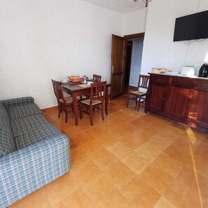 Een zithoek met een tafel en stoelen, een bank, een keukenblad met apparatuur en een deur naar een andere kamer.