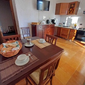 Een eetruimte met een houten tafel gedekt voor twee personen, een broodmand en een televisie boven het aanrecht.