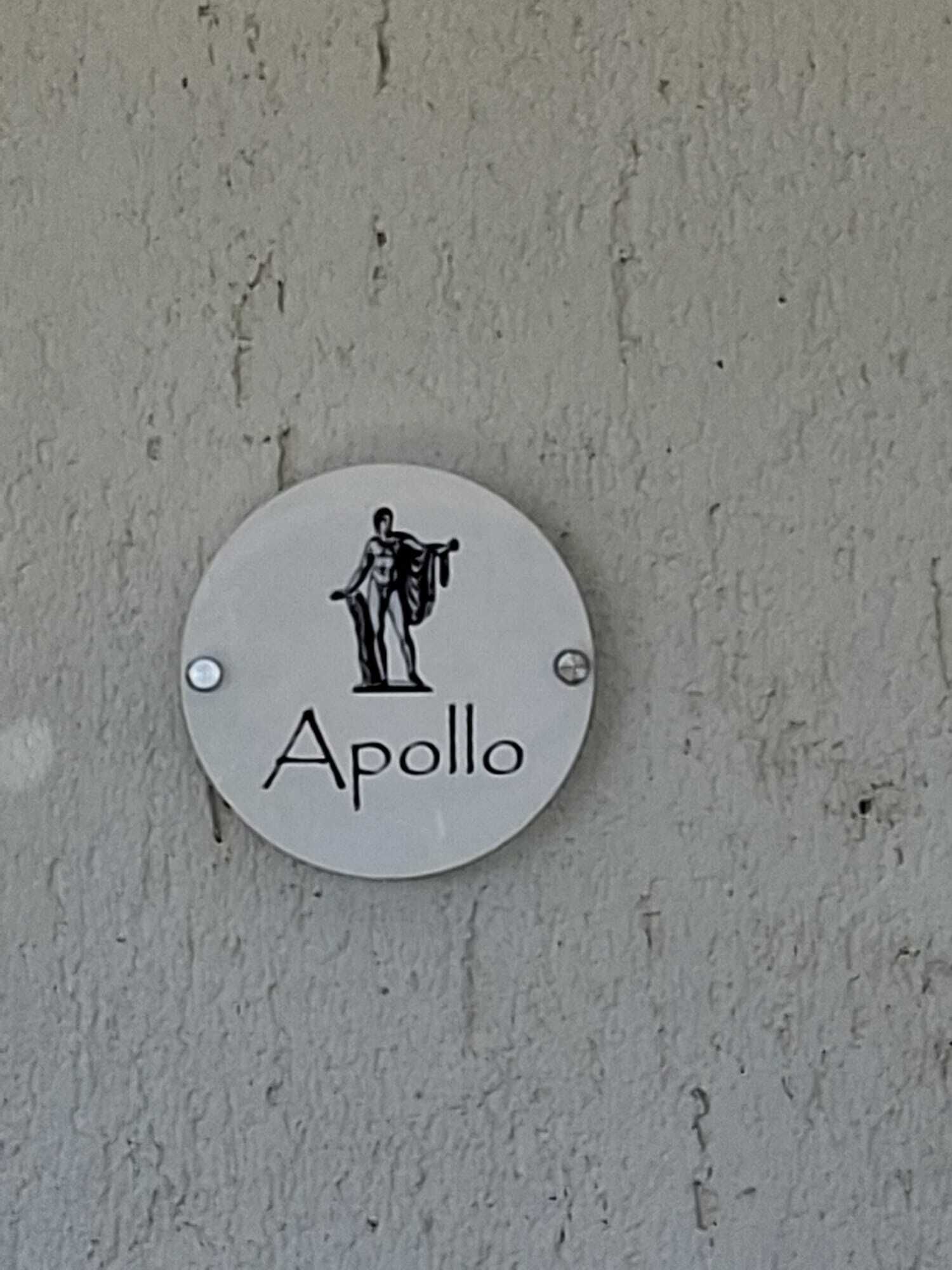 Ein rundes Schild mit einer Figur und dem Schriftzug 'Apollo' an einer strukturierten Wand.