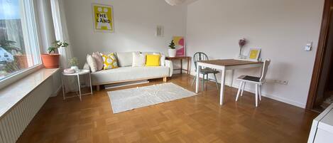 A modern living room with a sofa, a dining table, and chairs. There is a window with a view of trees, a radiator, and a potted plant.