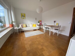 Uma sala moderna com sofá, mesa de jantar e cadeiras. Há uma janela com vista para árvores, um radiador e um vaso com planta.