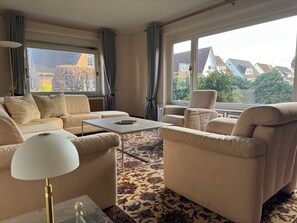 Un salon spacieux avec un canapé beige, des fauteuils, une table basse et une grande fenêtre donnant sur des maisons et des arbres.