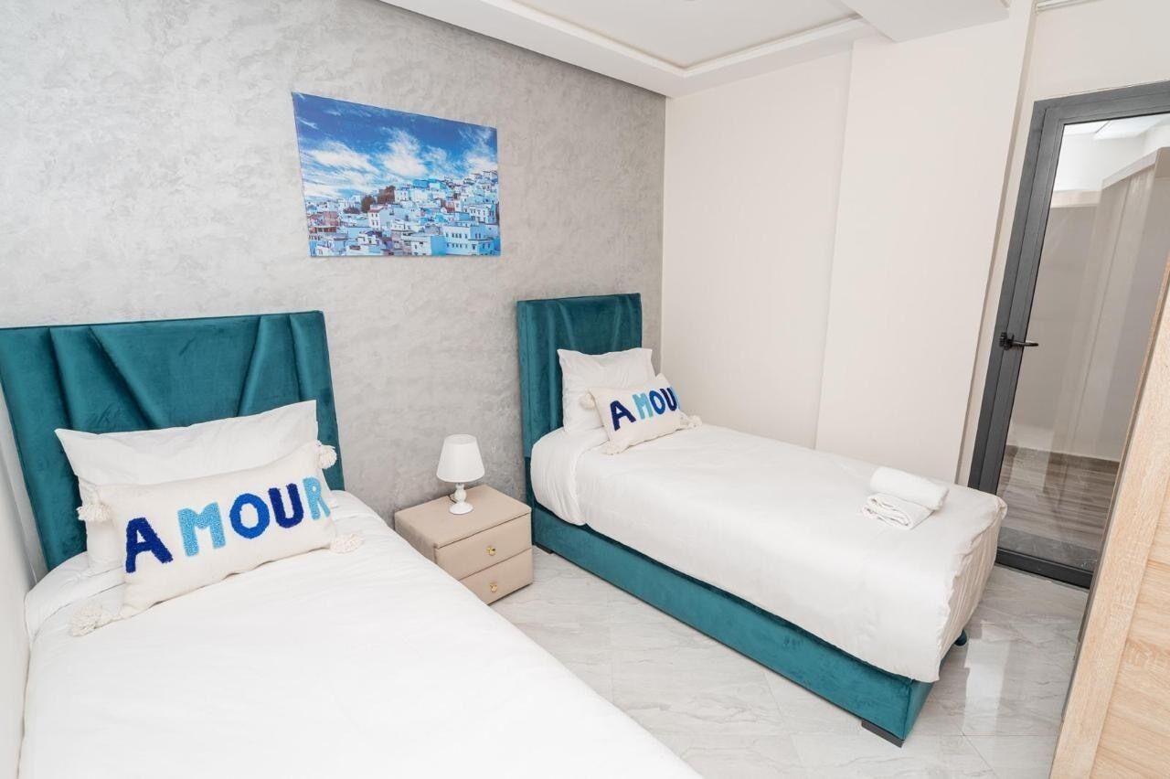 A hotel room with two beds, each with a teal headboard and white bedding. There is a nightstand with a lamp and a drawer, and a painting of a coastal town on the wall.