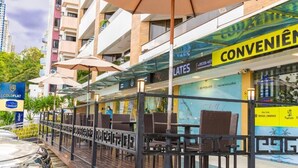 An outdoor seating area with umbrellas, a glass railing, and a view of a building with a 'CONVENIÊN' sign. - Flat 321-GF na praia de Cabo Branco (João Pessoa)