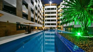 A swimming pool with lounge chairs and umbrellas, surrounded by a modern building with multiple floors and a palm tree. - Flat 321-GF na praia de Cabo Branco (João Pessoa)