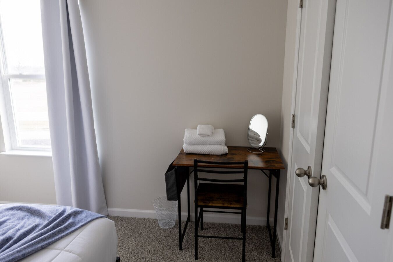 Ein kleines, ordentlich eingerichtetes Schlafzimmer mit einem Bett, einem Holztisch mit Stuhl, einem Spiegel und einer Tür.