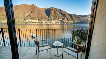 A balcony with two wicker chairs and a small table, overlooking a lake with mountains in the background.