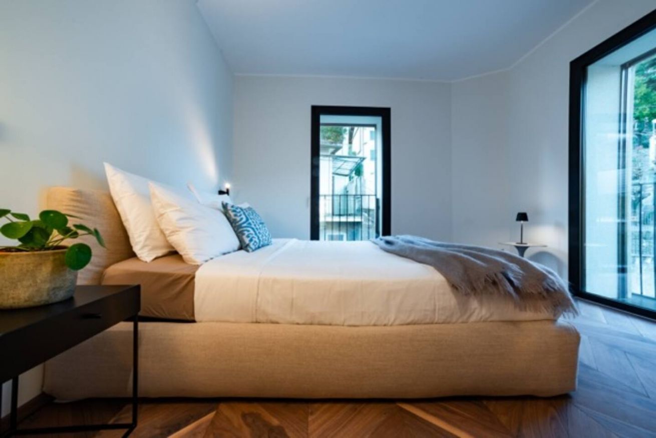 Ein modernes Schlafzimmer mit einem großen Bett, einem kleinen Tisch mit Pflanze und Blick durch ein Fenster.