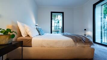 Ein modernes Schlafzimmer mit einem großen Bett, einem kleinen Tisch mit Pflanze und Blick durch ein Fenster.